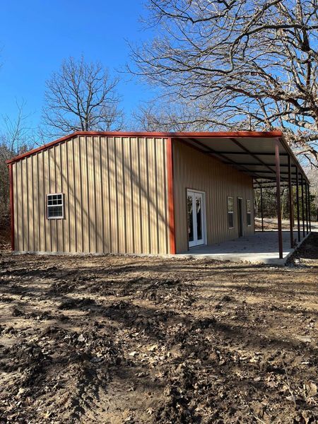 Tan metal building with a covered patio, set in a dirt yard with trees and blue sky.