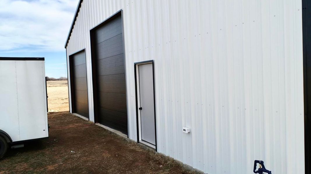 White metal building with black garage doors, small white door, and a white trailer.