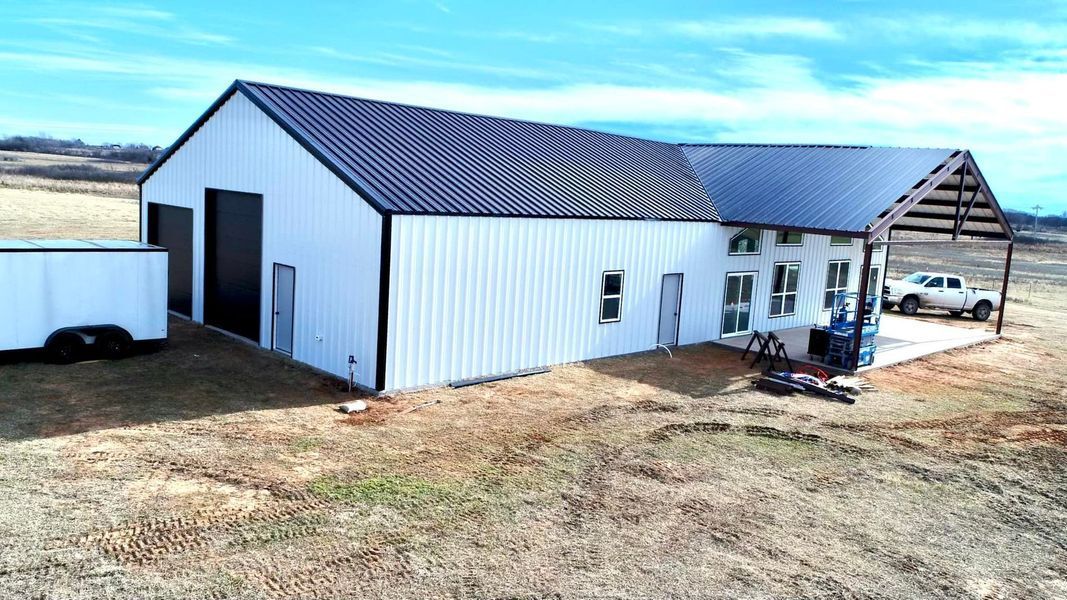 White metal building with dark roof and attached open-air awning; trailer and pickup truck nearby.