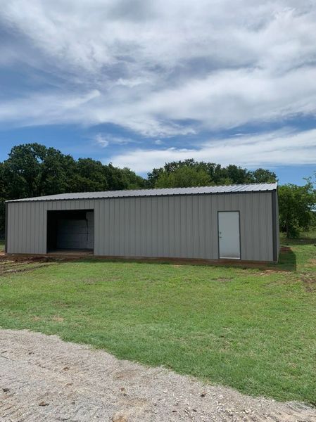 Metal shed with open bay and white door on a grassy field under a cloudy sky.