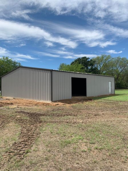 Gray metal barn with open front and a door, set in a grassy field under a cloudy blue sky.