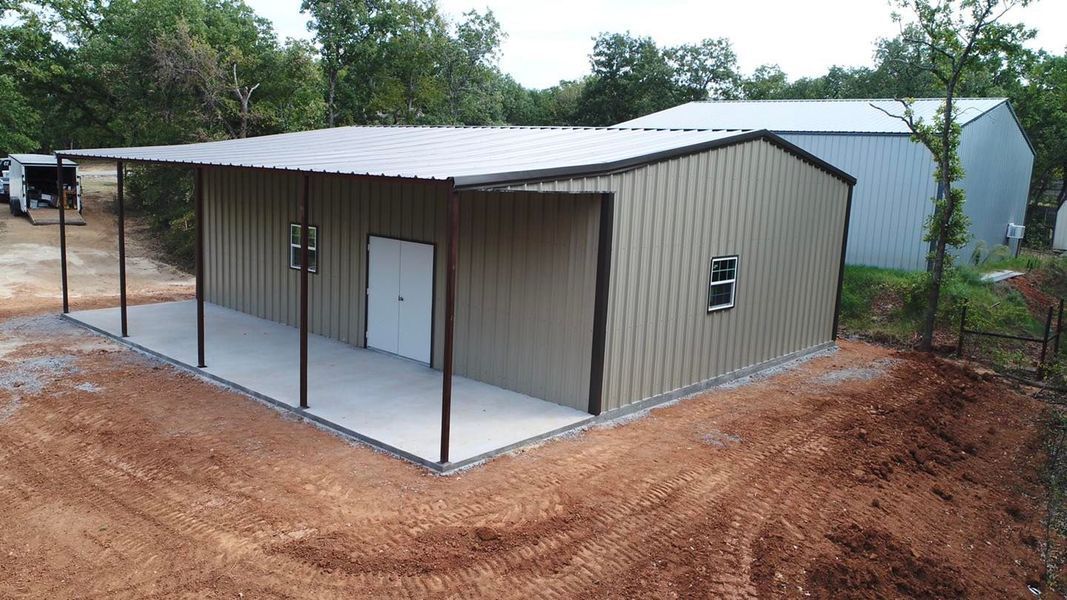 Metal building with brown trim and covered porch on a dirt lot. Another building in background.