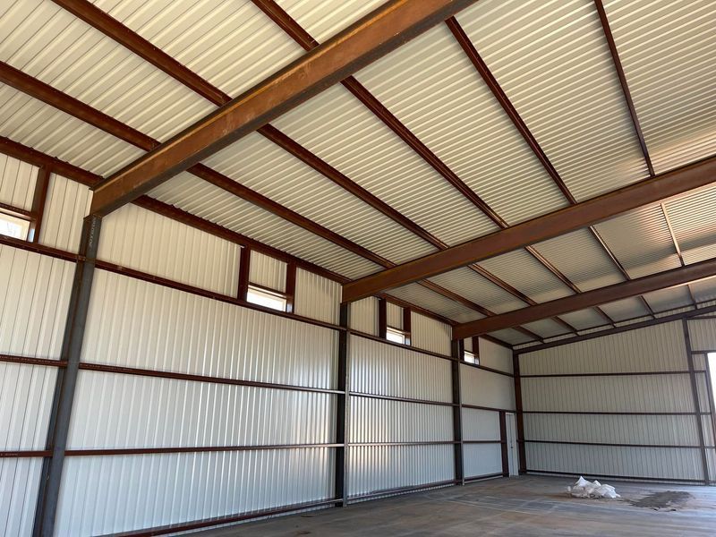 Interior of a metal building with white corrugated walls and ceiling, brown beams, and small windows.