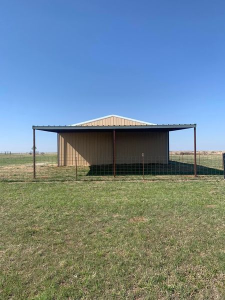 Metal-roofed barn with an open-sided shelter in front, on a grassy field under a blue sky.