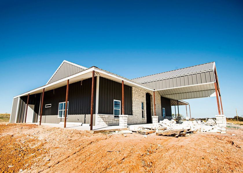 New house under construction with a metal roof and dark siding, on a brown dirt lot under a blue sky.