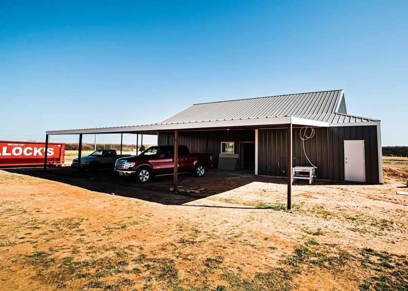 Red pickup truck and car parked under a metal carport next to a brown barn in a dry, outdoor setting.
