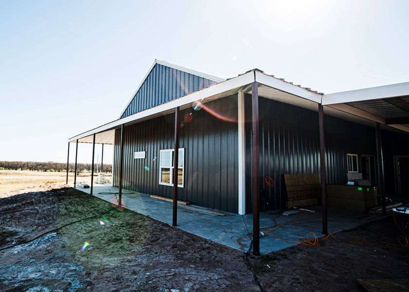 Dark gray metal building with white trim, a porch, and a gravel yard under a blue sky.