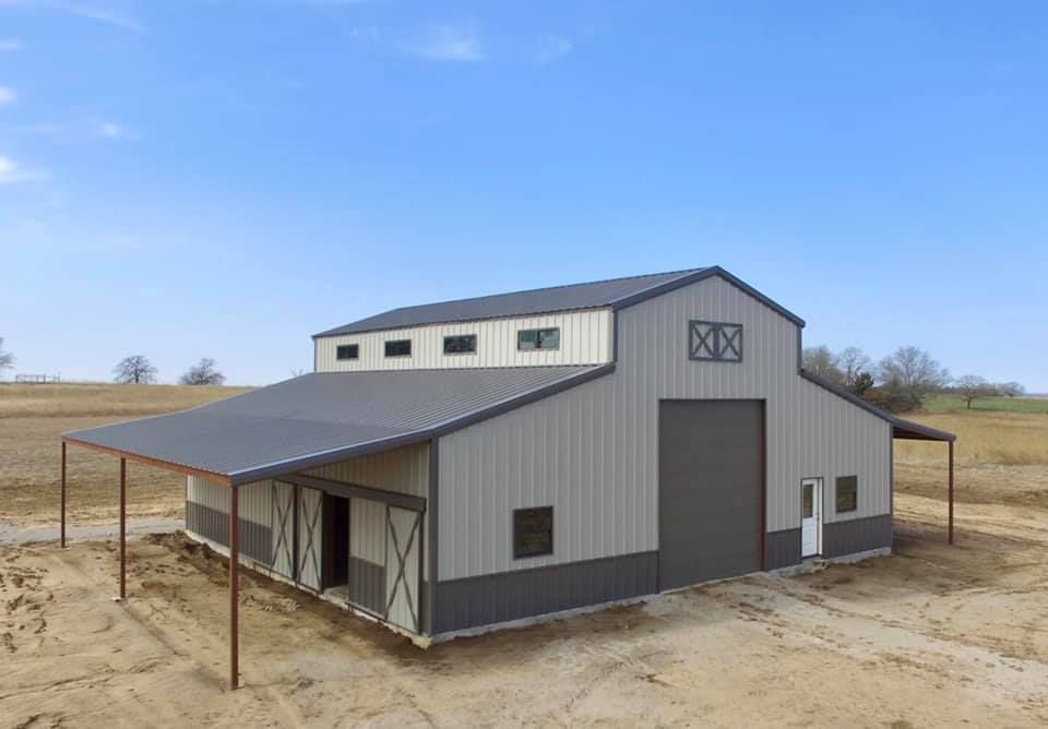 Two-story metal barn with gray roof and siding, brown trim, and a covered porch, set on a field under a blue sky.