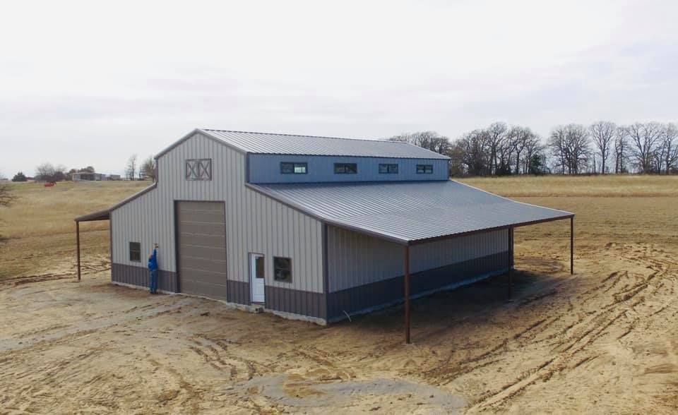 Metal barn with gray roof and blue accents in a field. Man standing by a large garage door.
