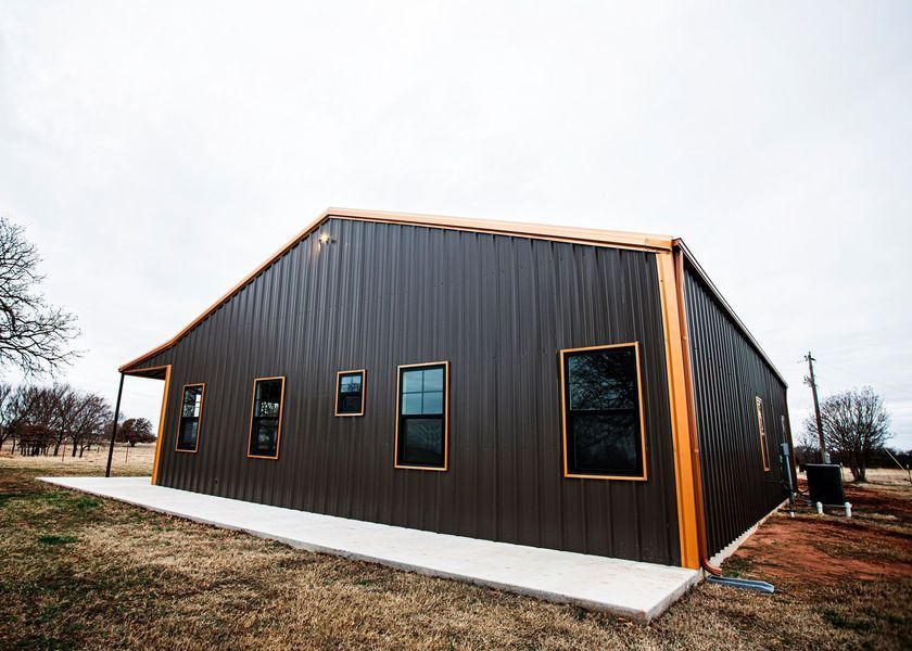 Brown metal building with dark windows and orange trim on a concrete slab in a field under a cloudy sky.
