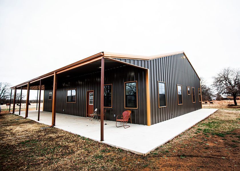 Brown metal-sided house with a porch and concrete patio on a grassy lot under an overcast sky.