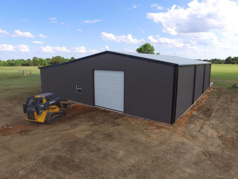 Dark gray metal building with a large white garage door; yellow skid steer in the foreground.