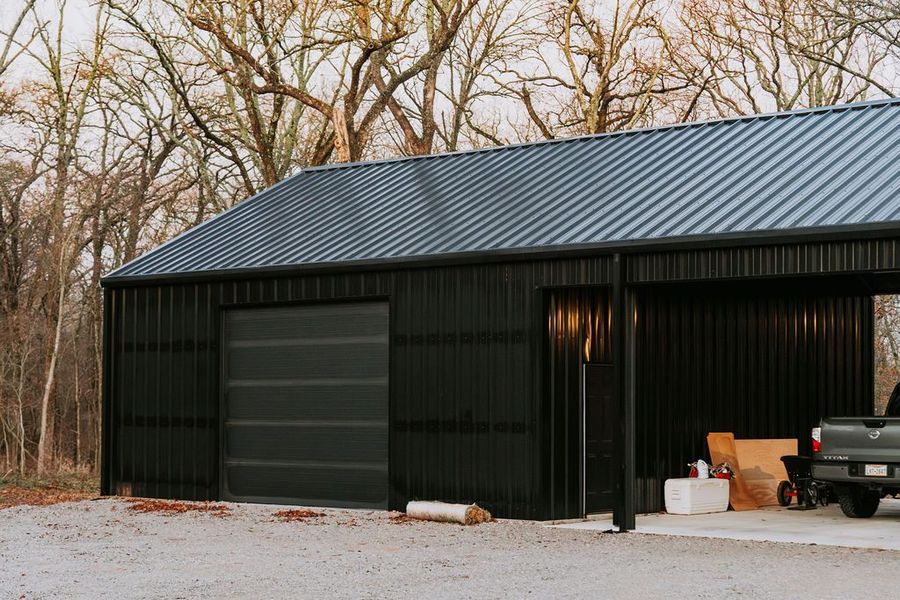 Black metal shed with a gray garage door and carport, set in a wooded area.