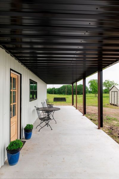 Covered porch with white building, black roof, and chairs. Open to green field.