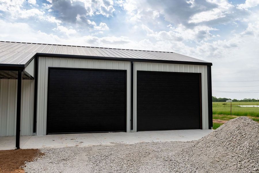 Two-car garage with black doors, white siding, and a silver metal roof. Gravel driveway and a pile of gravel nearby.