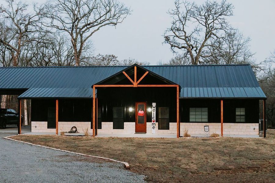 Black-sided ranch-style house with orange support beams and a dark metal roof. Red front door, light-colored brick base.
