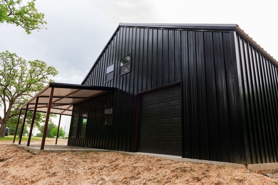 Black metal-clad building with carport and garage door on a dirt lot, under an overcast sky.