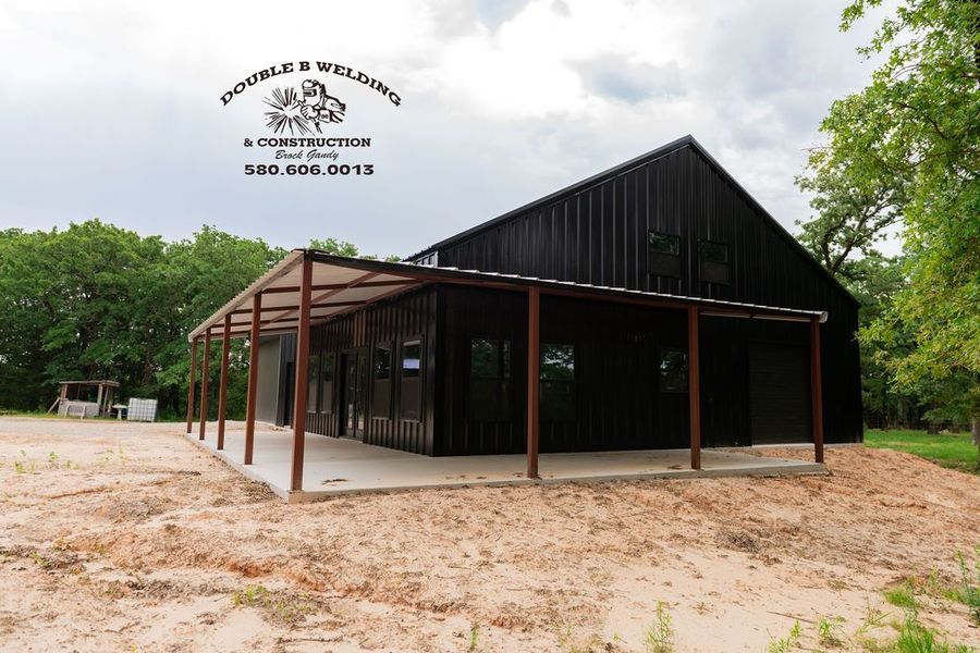 Black barn with a covered porch on a dirt lot, surrounded by trees under a cloudy sky.