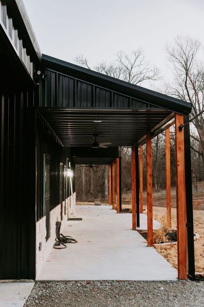 Black and brown modern porch with concrete walkway, wood columns, and metal roof. Trees in the background.
