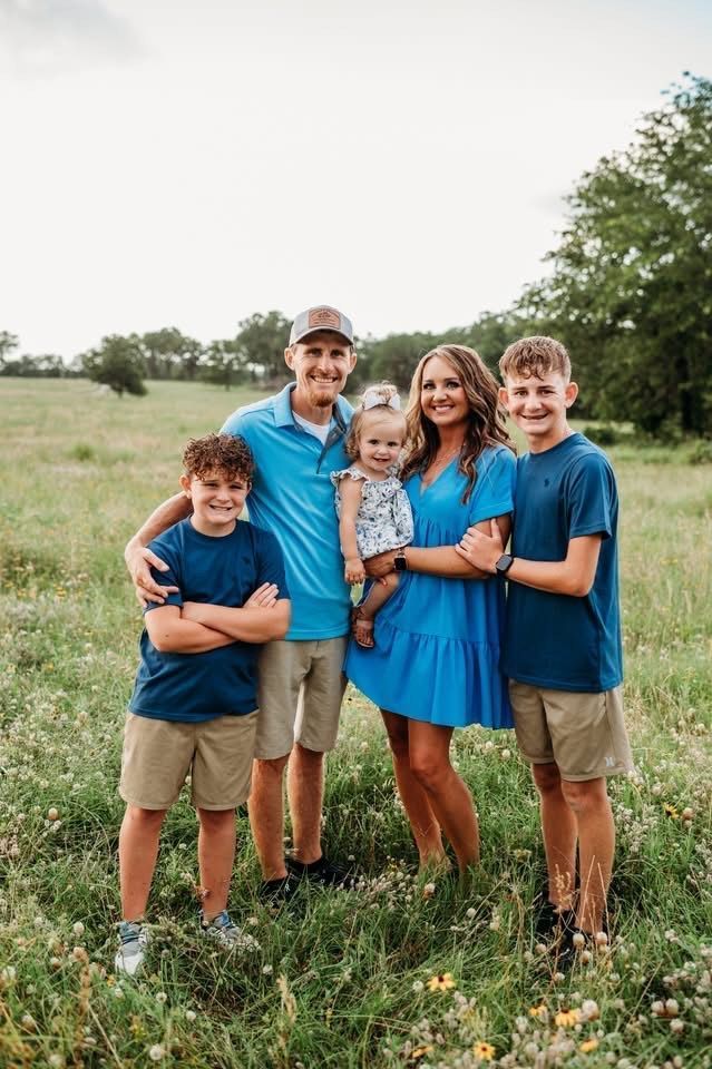 Family of five posing in a grassy field. Mother in blue dress holds baby. Older boys wear blue shirts and khaki shorts.