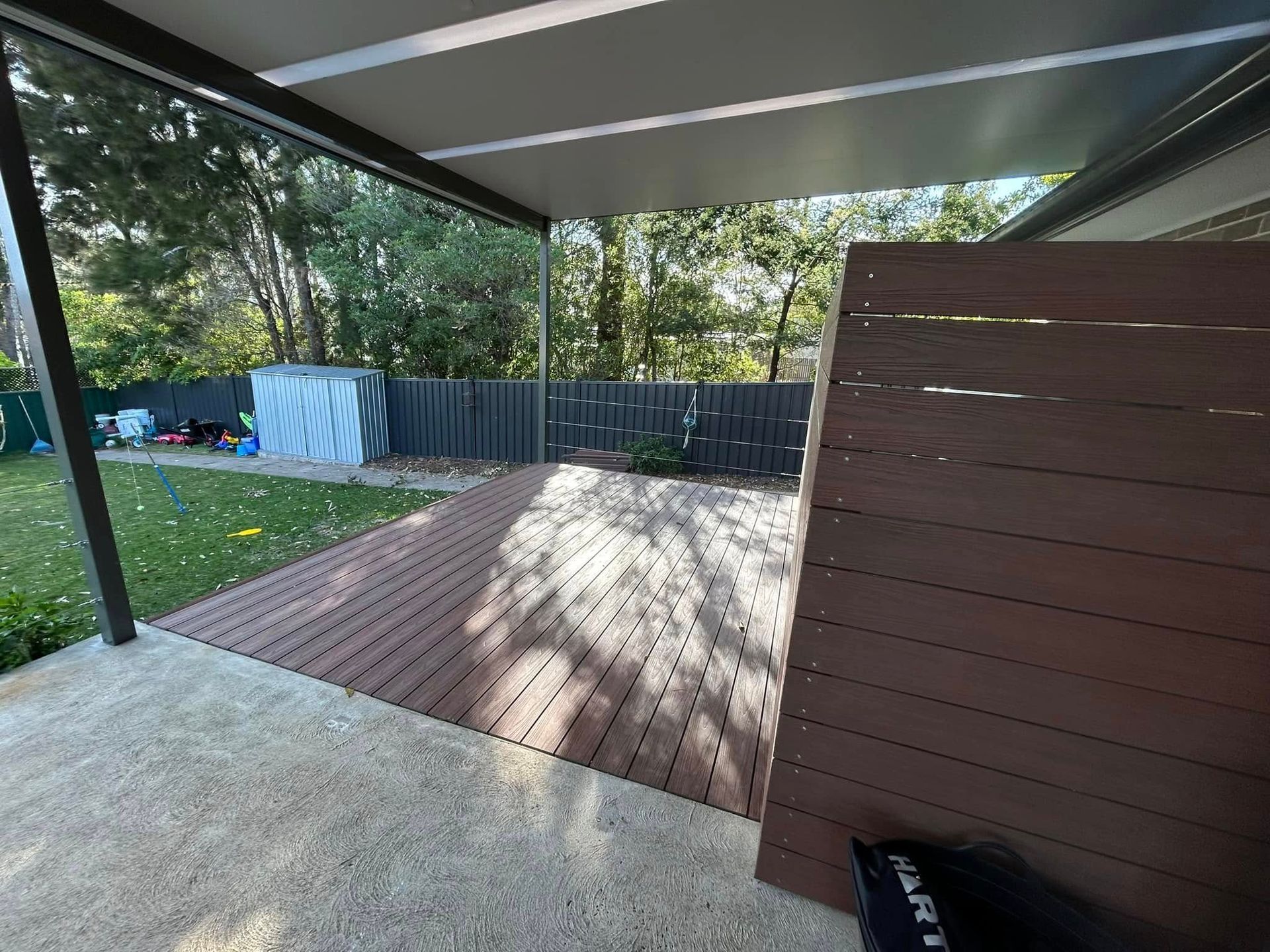 A wooden deck under a covered patio with a fence in the background.