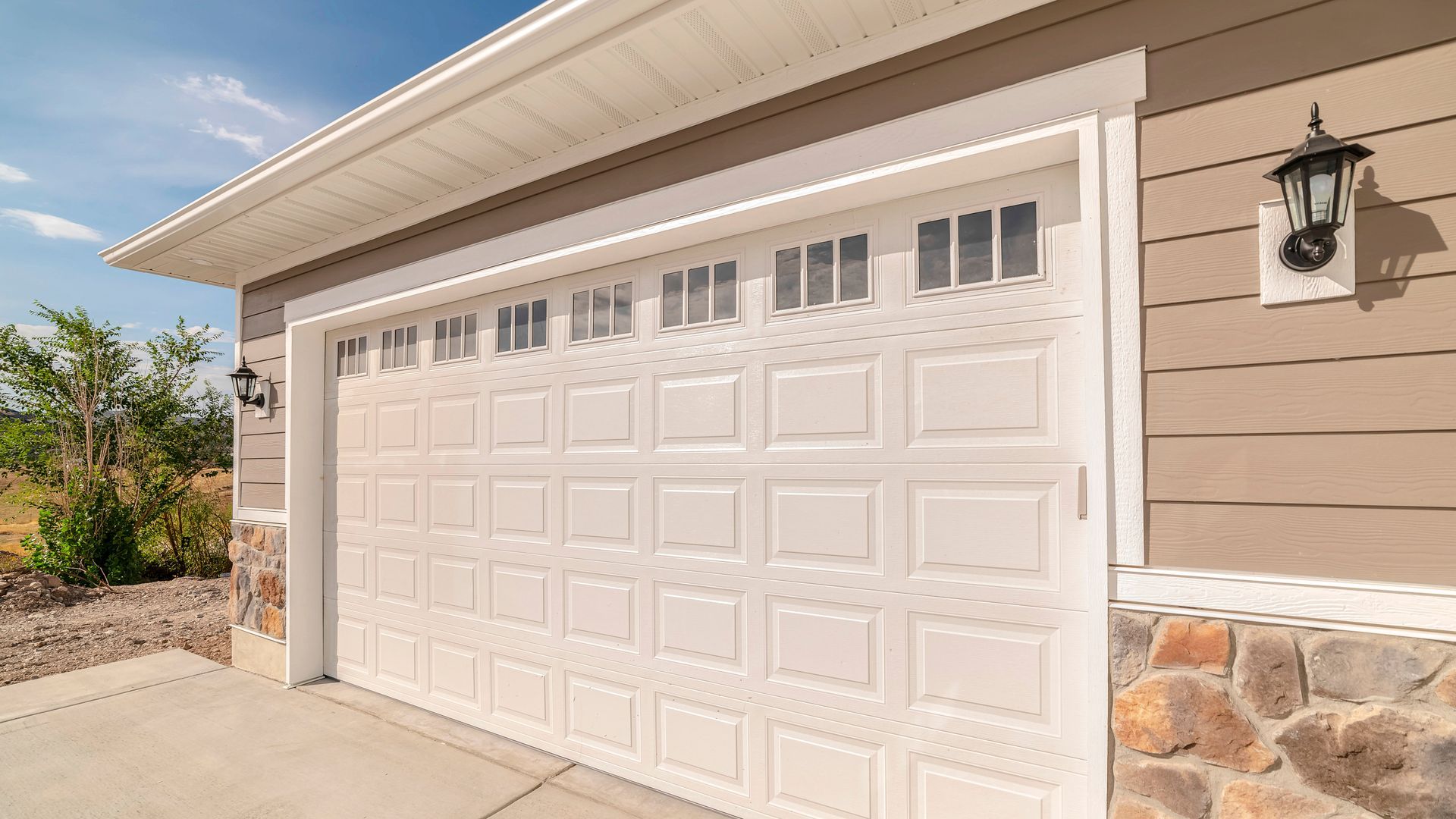 White paneled garage door on a beige suburban house with stone trim and a wall lantern