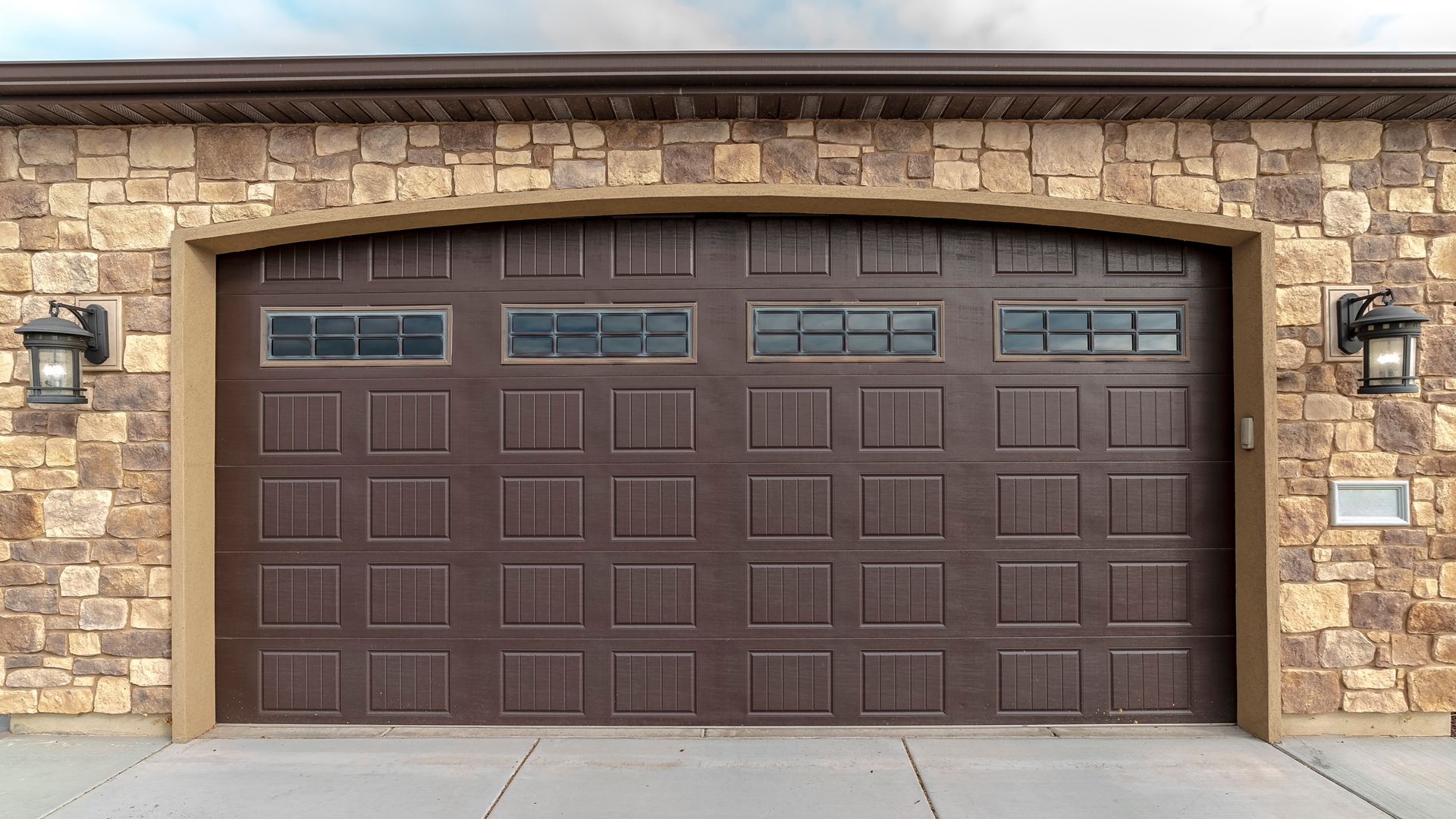 Brown two-car garage door in a stone house facade with decorative windows and wall lanterns