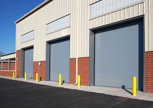 A beige industrial building with three gray roll-up bay doors, red brick bases, and yellow bollards in a parking lot.