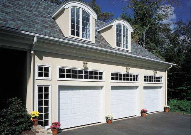 A three-car garage with white doors, transom windows, and two arched dormers on a shingled roof set against trees.