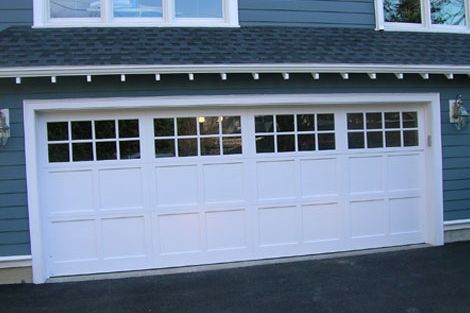 White, four-panel garage door with top window panes, set in a blue-sided house with a shingled roof.