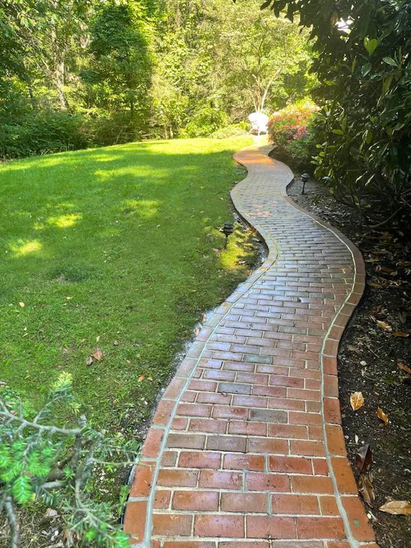 Brick pathway curves through a green lawn towards a sunlit, treed area.