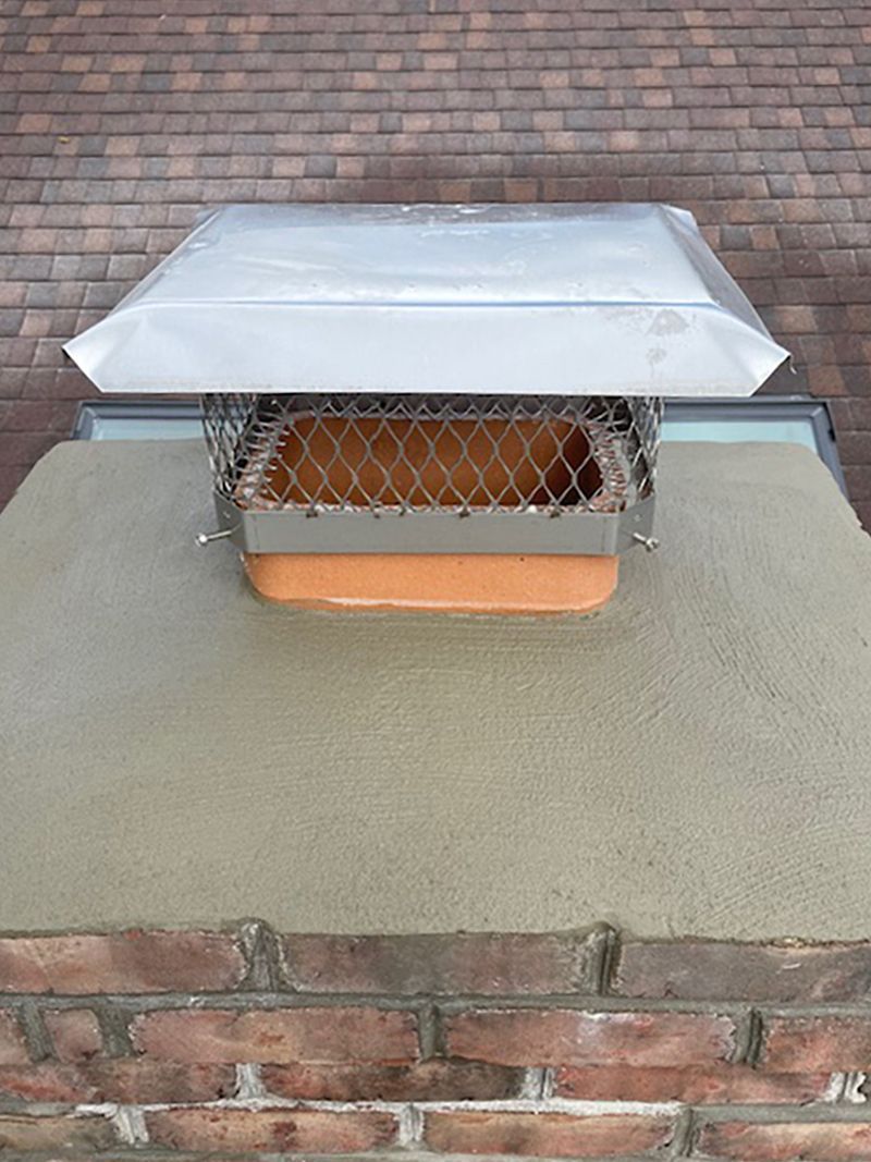 Chimney with metal cap and screen on a brick structure, viewed from above against a brown shingle roof.