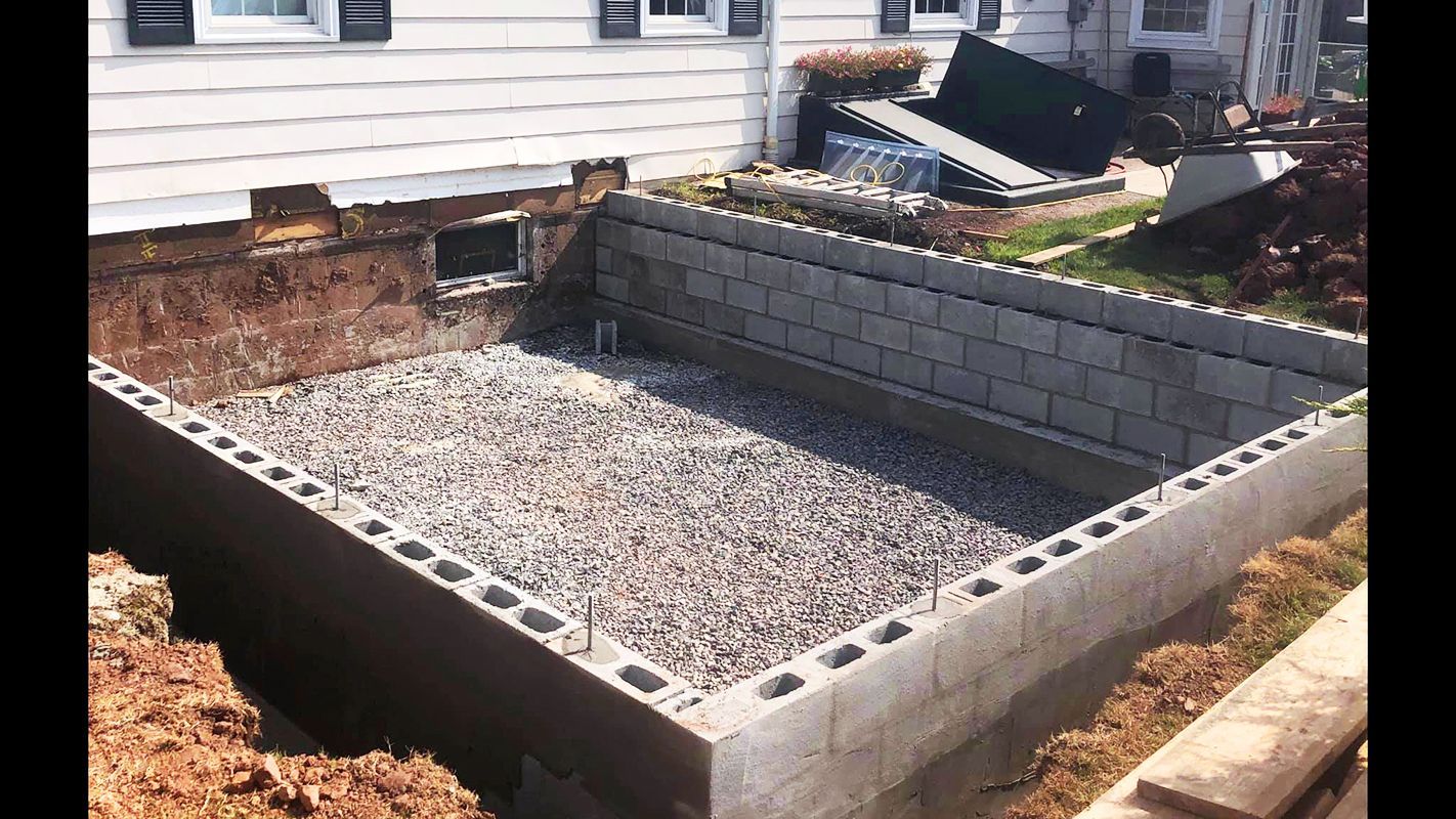 Construction site: rectangular foundation excavation with cinder block walls, gravel base, next to a house.