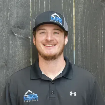 A man wearing a hat and a polo shirt is smiling while standing in front of a wooden fence.