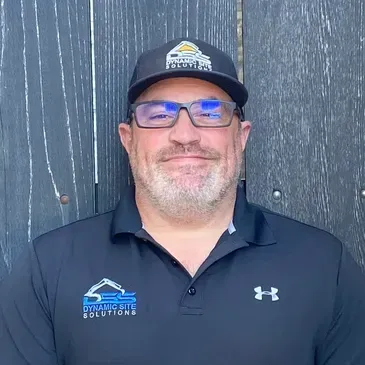 A man wearing a hat and glasses is standing in front of a wooden fence.