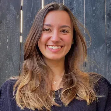 A woman with long hair is smiling in front of a wooden fence.