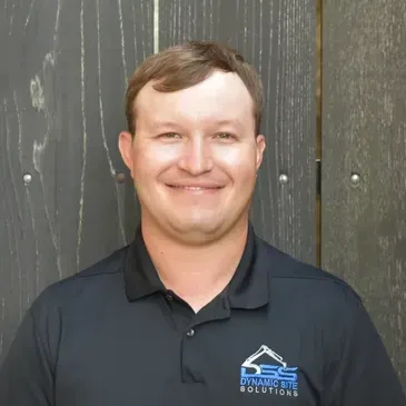 A man in a black shirt is smiling in front of a wooden wall.