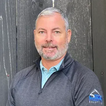 A man with a beard is standing in front of a wooden wall.