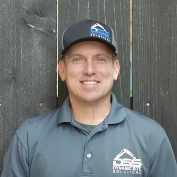 A man wearing a hat and a polo shirt is smiling in front of a wooden fence.