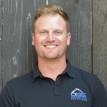 A man in a black polo shirt is smiling in front of a wooden wall.