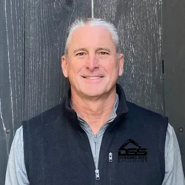 A man wearing a black vest is standing in front of a wooden wall.