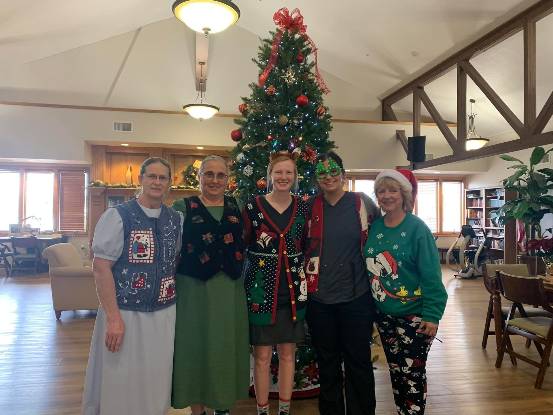 Five people in festive sweaters pose by a decorated Christmas tree in a room with wood beams.