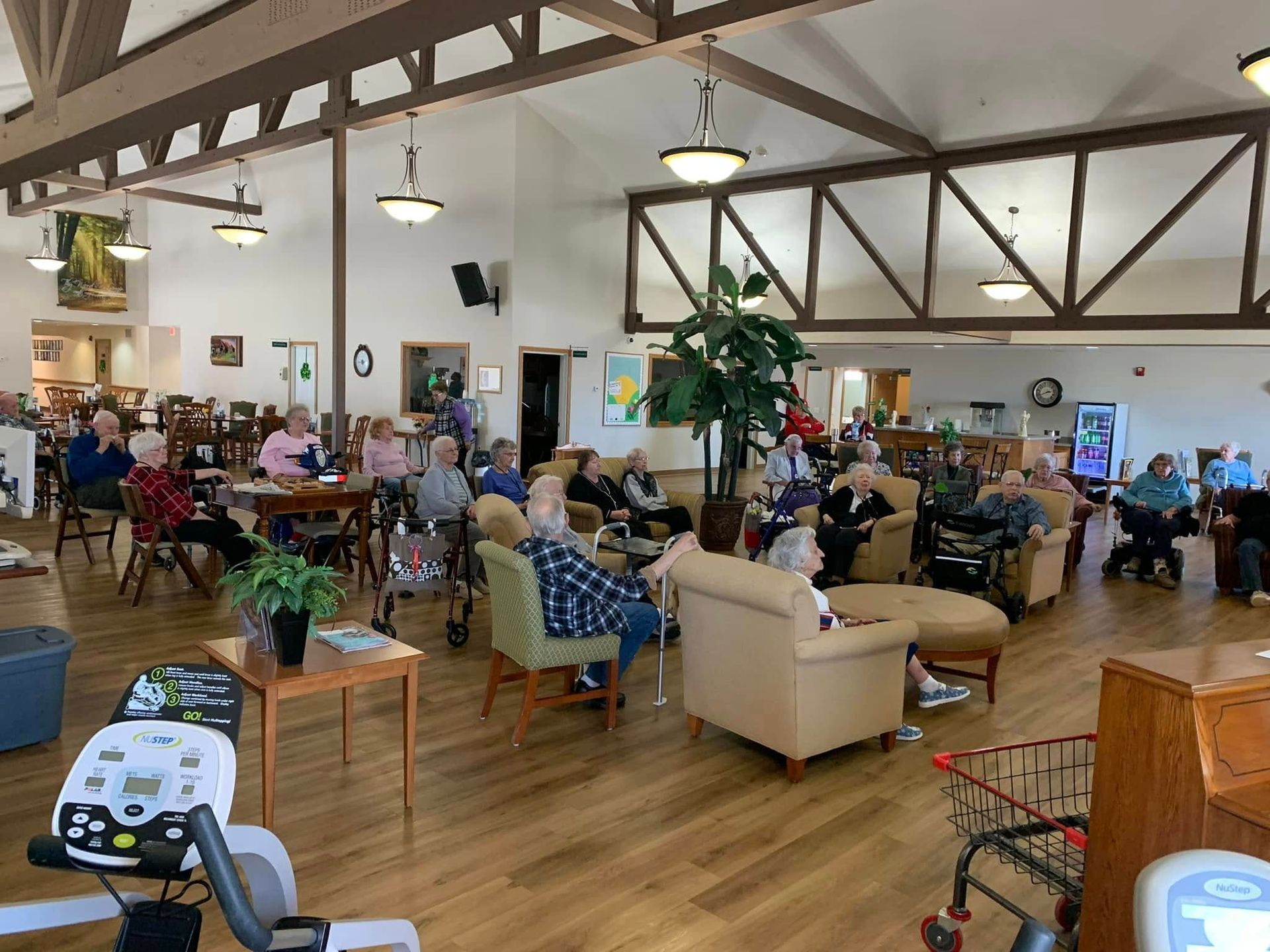 Seniors in a large community room socializing; some sitting, others with walkers. Wood floor, high ceiling.