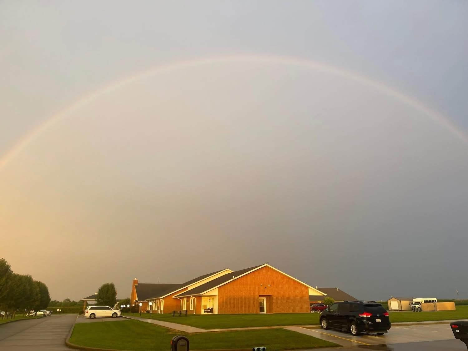 Rainbow arches over a residential area with a brick building and parked cars.