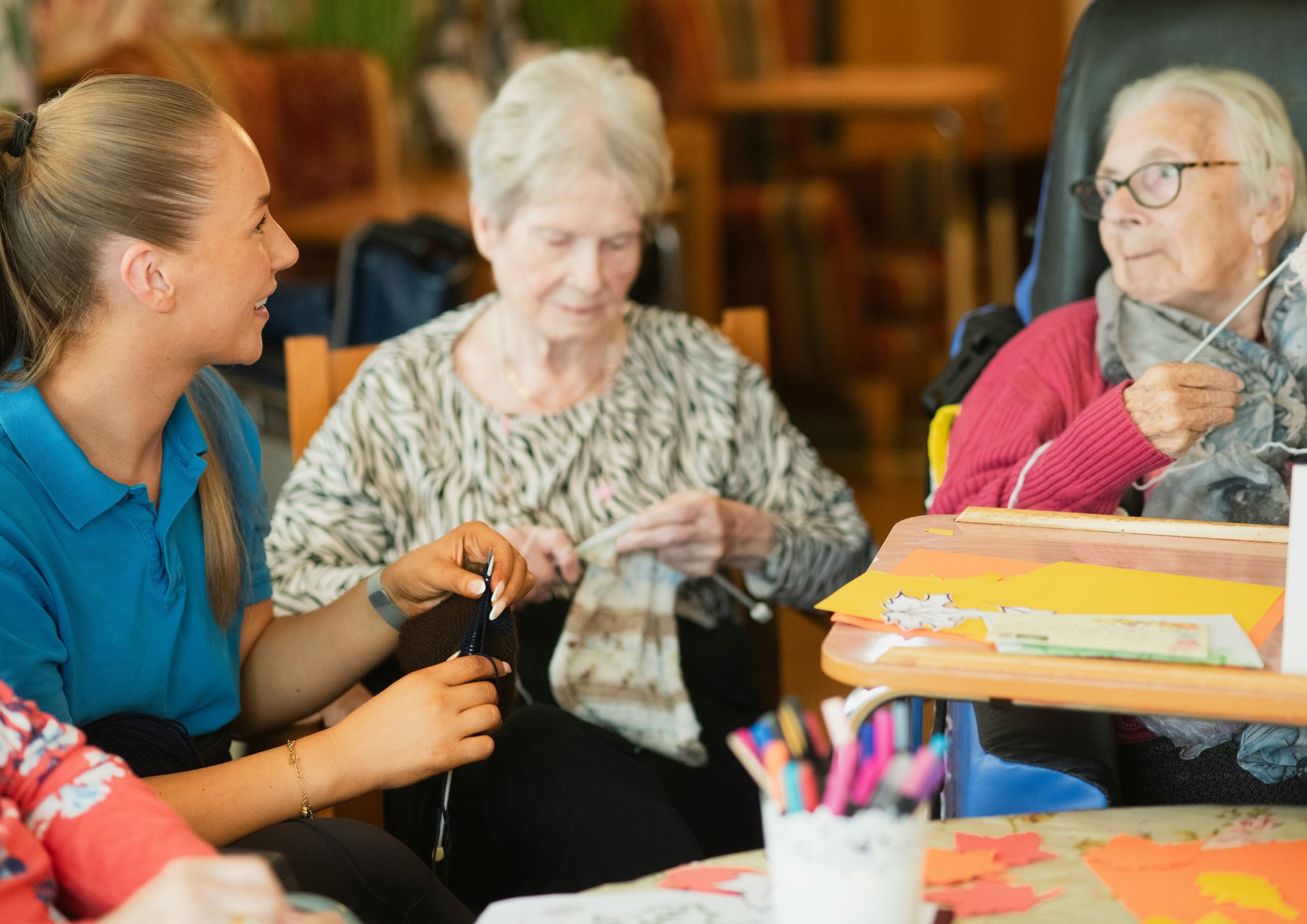 Woman in blue shirt conversing with two elderly women; one knits, the other crafts. Indoor setting, colorful details.
