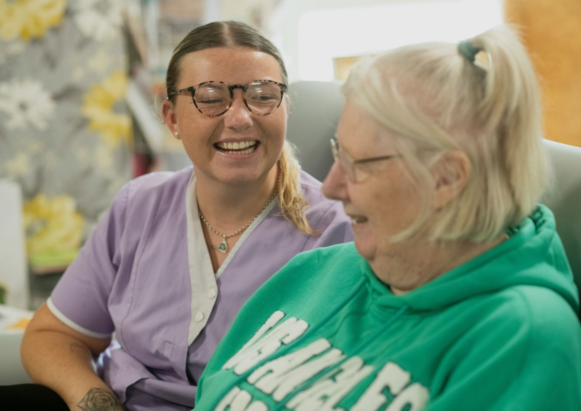 Caregiver in lavender uniform smiles at older person in green hoodie. They sit indoors.