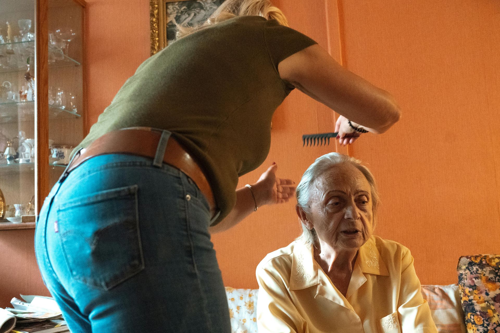 Woman cutting elderly person's hair indoors, using comb and scissors. Orange wall background.