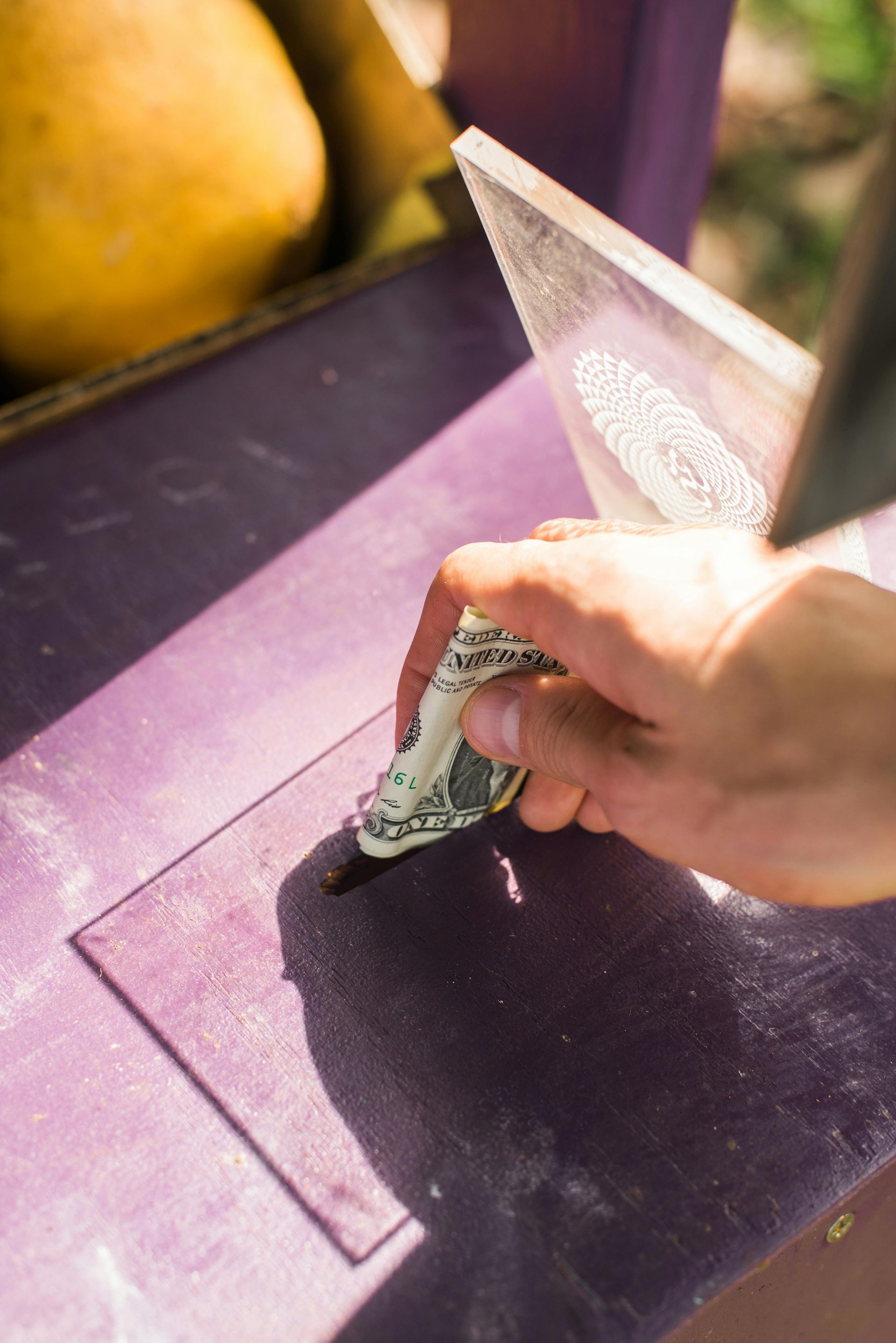 Hand inserting a dollar bill into a purple donation box with a transparent lid, possibly for fruit.