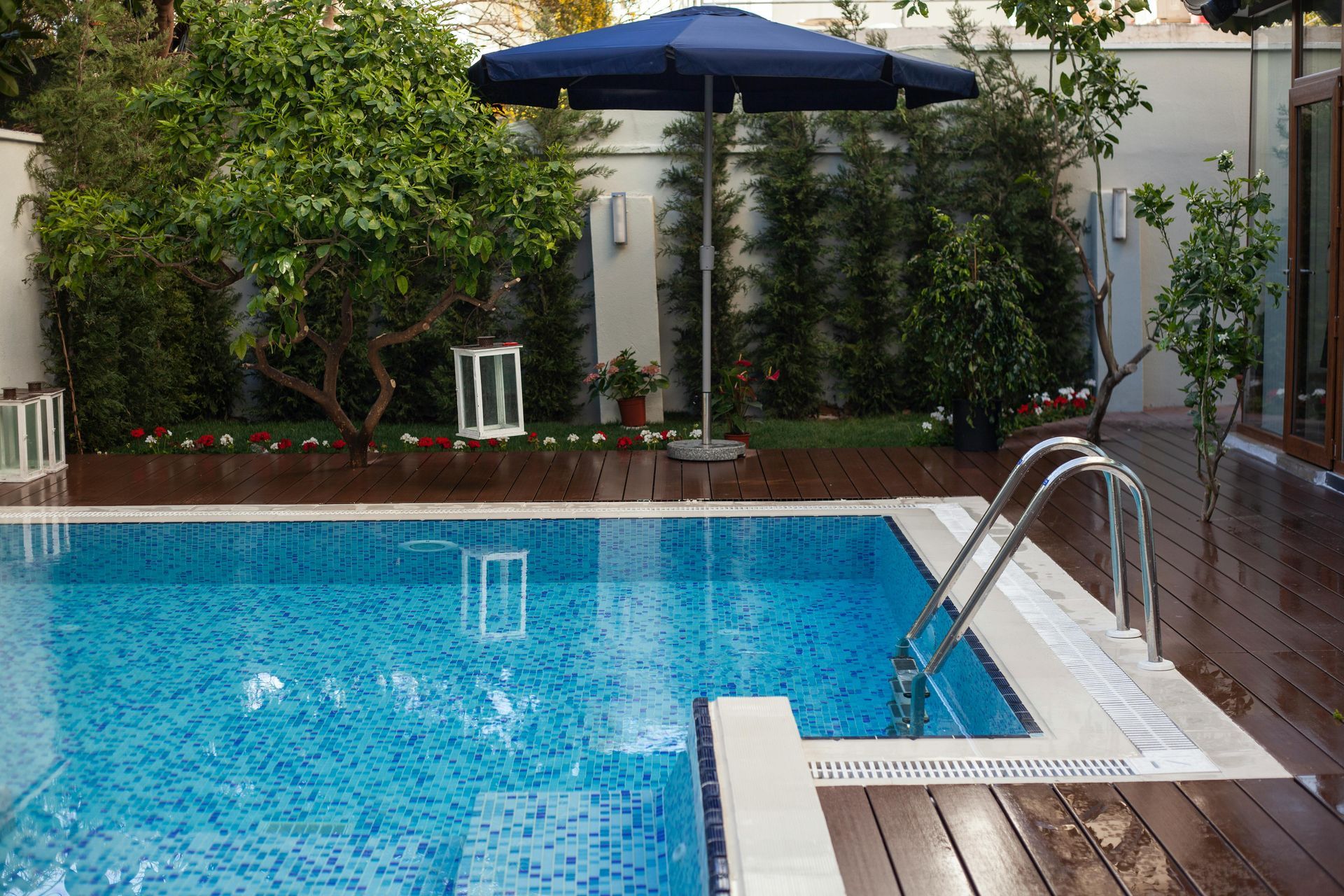 Blue swimming pool with ladder, patio chairs, and umbrella in a garden setting