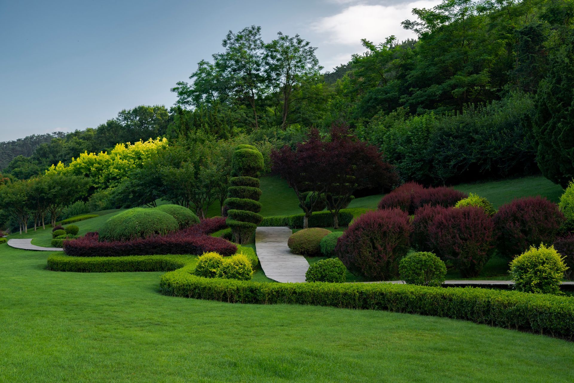 Lush green lawn, manicured bushes, and a pathway lead up a hillside with colorful plants and trees.
